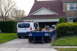 Low blue dumpster full of cardboard and construction debris in front of a full garage and next to a white worker's van