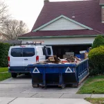 Low blue dumpster full of cardboard and construction debris in front of a full garage and next to a white worker's van
